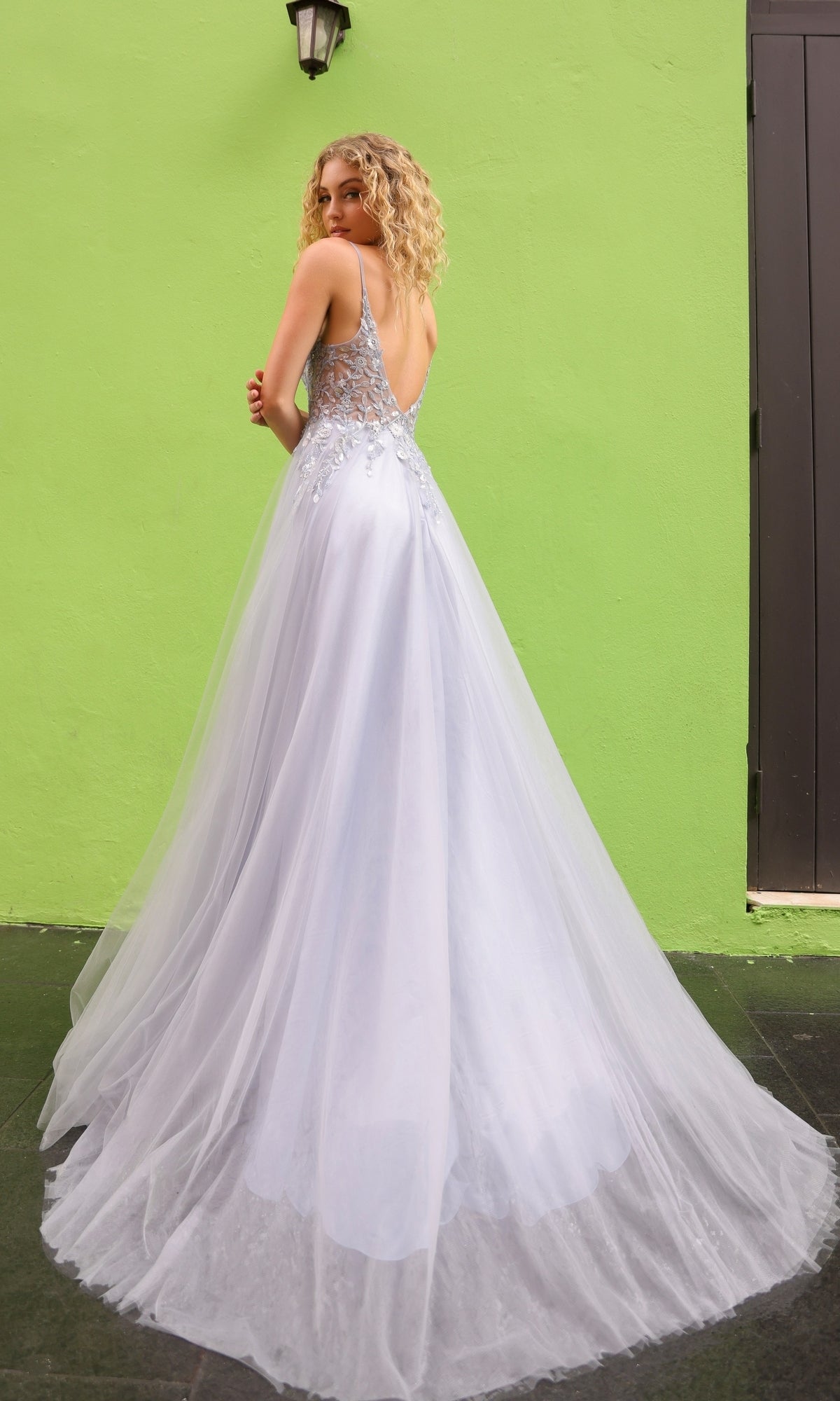 A woman with curly blonde hair wears the Deep V-Back Long Tulle A-Line Prom Dress Q1391 in light blue, featuring a backless design and floor-length skirt, while standing before a bright green wall.